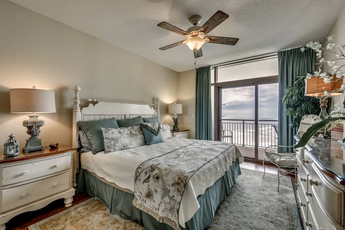 A serene bedroom with a comfortable bed adorned with decorative bedding, a large window offering a view, a dresser topped with a lamp, and tranquil accents throughout. This space radiates a sense of peaceful retreat, perfect for relaxing or reading after a day at the beach.