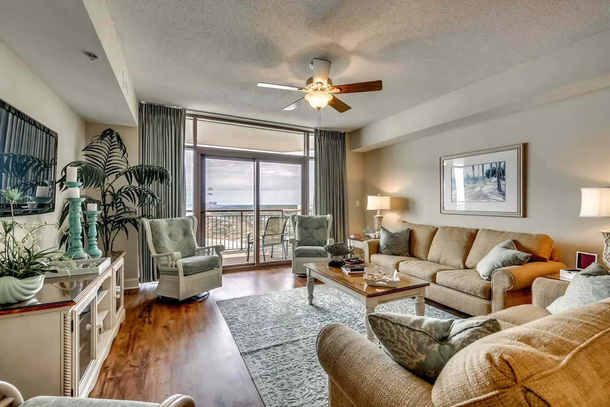 A cozy living room corner with a sofa, coffee table, and a window view looking toward the balcony. The scene highlights comfort and family-friendly amenities, with space for board games, reading, and enjoying the sunset through the window. Decorative accents like plants and a ceiling fan create a welcoming mood.
