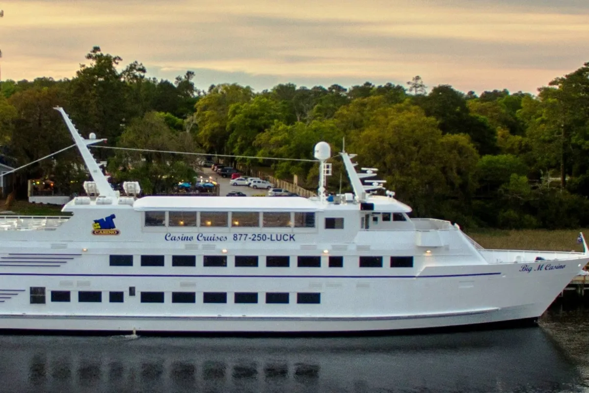 The image captures a large casino boat docked along a scenic river, surrounded by lush trees and offering views of the expansive waterway. The setting is vibrant, with a hint of adventure and the promise of unique entertainment and relaxation on the water.