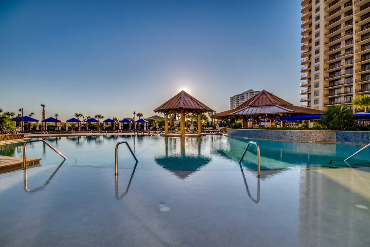A serene sunset view of a swimming pool area framed by tall palm trees and tropical cabanas. The tranquil water reflects the orange and pink hues of the sky, inviting guests to relax, socialize, or enjoy a refreshing swim as the day winds down.