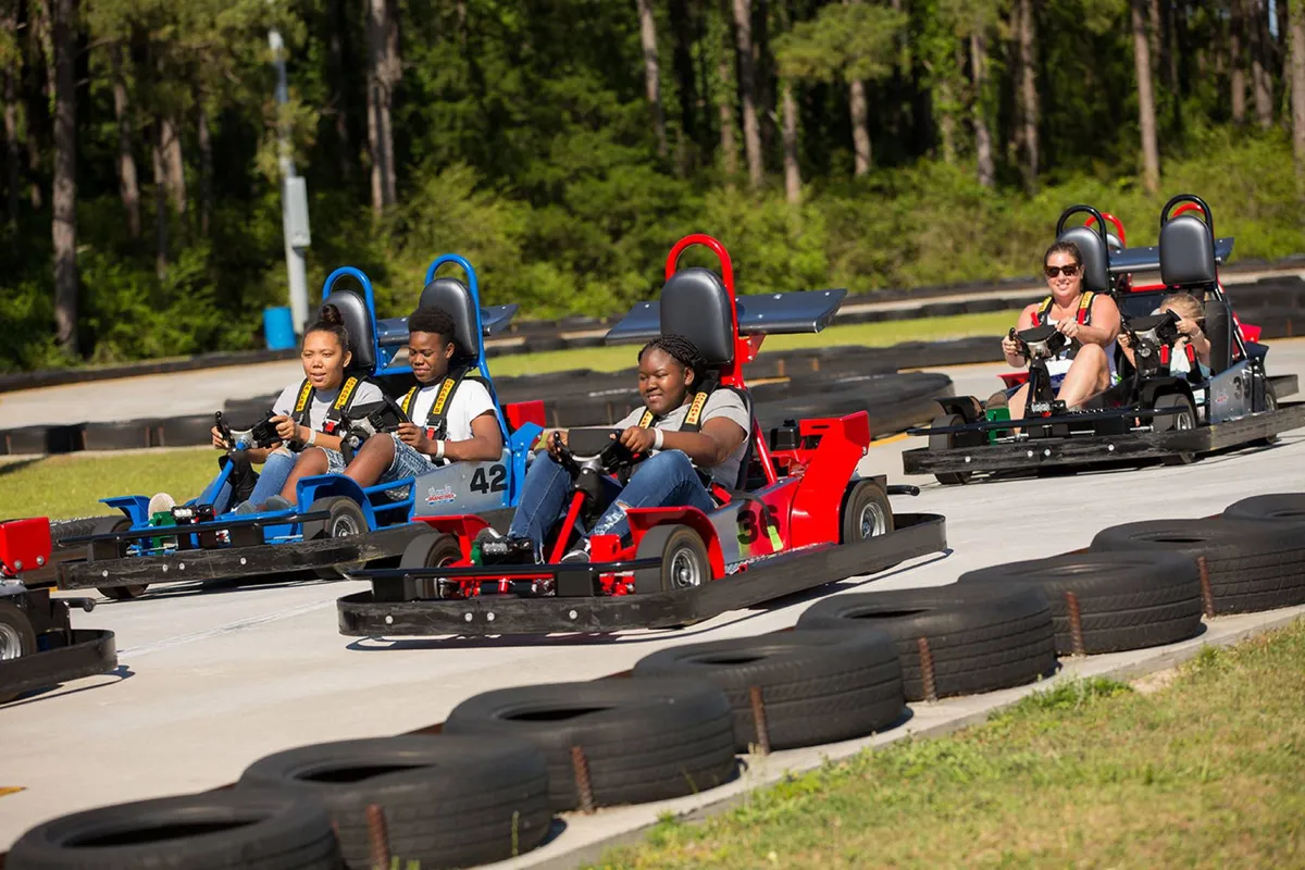 An energetic exterior scene at a go-kart track, with several participants racing and cheering. The playful mood conveys family fun and friendly competition, with visible excitement among groups.