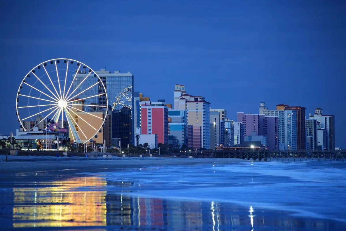 This vibrant exterior scene presents a ferris wheel illuminated against a backdrop of colorful buildings, with ocean reflections adding to the playful, festive mood. The setting suggests a classic beachfront amusement area bustling with families and visitors.