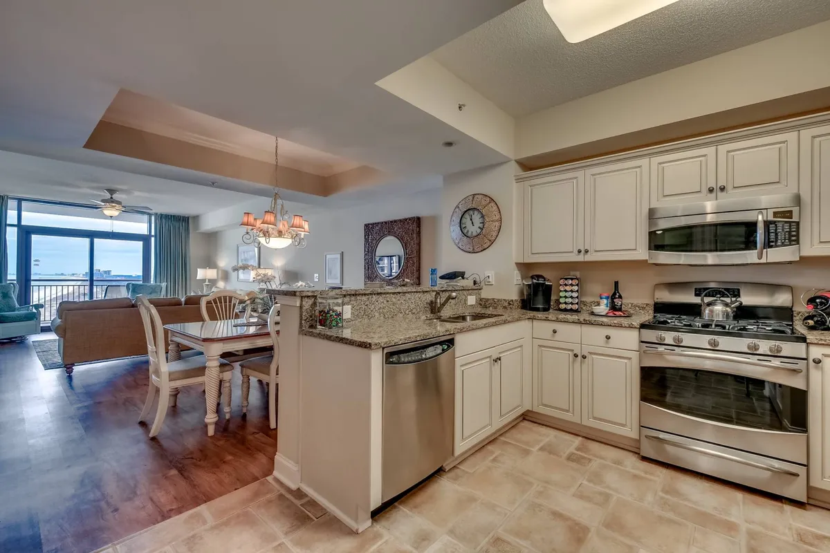 A wide shot encompassing the living area and dining space, with a granite countertop and stainless steel appliances in the background. The living room includes a sofa and large windows overlooking the ocean, highlighting both comfort and openness. The space is clearly designed for socializing, meals, and entertaining.
