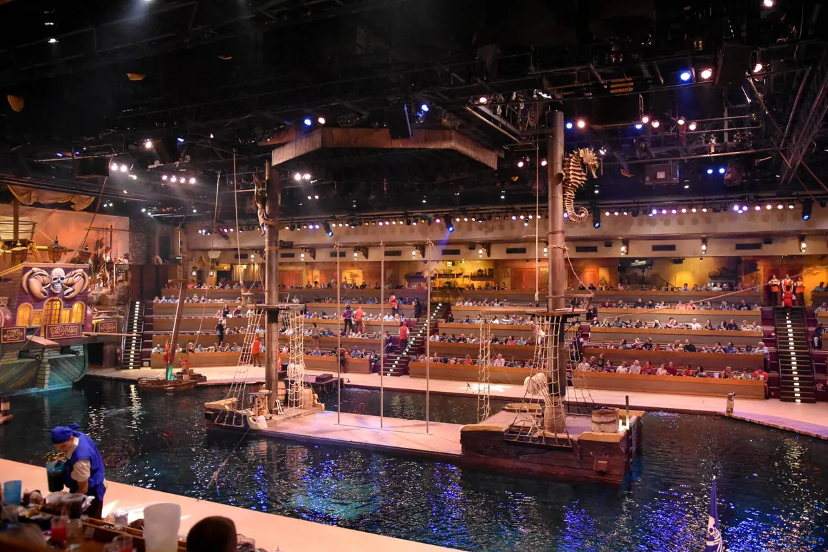 A lively interior scene showing a theater with a watery stage, audience seating, nautical-themed decorations, and dramatic lighting—setting the stage for an immersive, family-friendly performance.