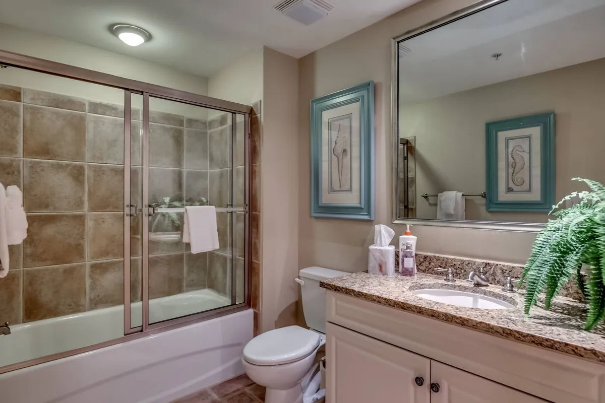 A luxurious bathroom boasting a glass shower enclosure, granite countertop, stylish framed artwork, and a leafy plant accent. The atmosphere is tranquil, inviting self-care and relaxation.