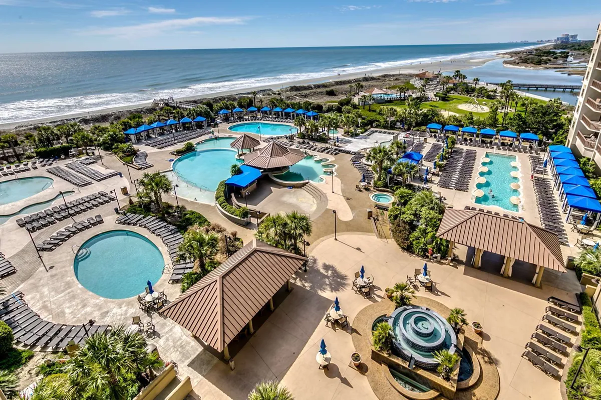 A vibrant exterior scene featuring expansive swimming pools surrounded by palm trees, lounge chairs, and tiki huts. The setting is lively and inviting, with the blue ocean visible in the background, and plenty of space for sunbathing, swimming, and relaxing by the water.