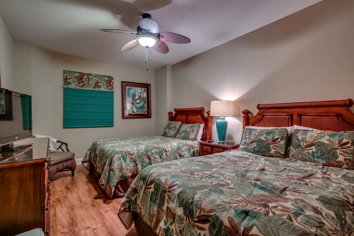 A spacious guest bedroom featuring two large beds with floral bedding, a bedside lamp, a colorful piece of art, a dresser with a mirror, and a comfortable chair. The room feels welcoming and ideal for families or friends sharing a space.