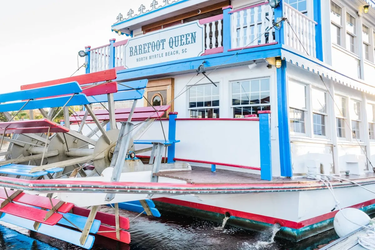 A vibrant exterior shot of the Barefoot Queen Riverboat docked by the water, featuring its classic paddle wheel and colorful facade. The atmosphere is lively, suggesting group dining, sightseeing tours, and social gatherings on board.